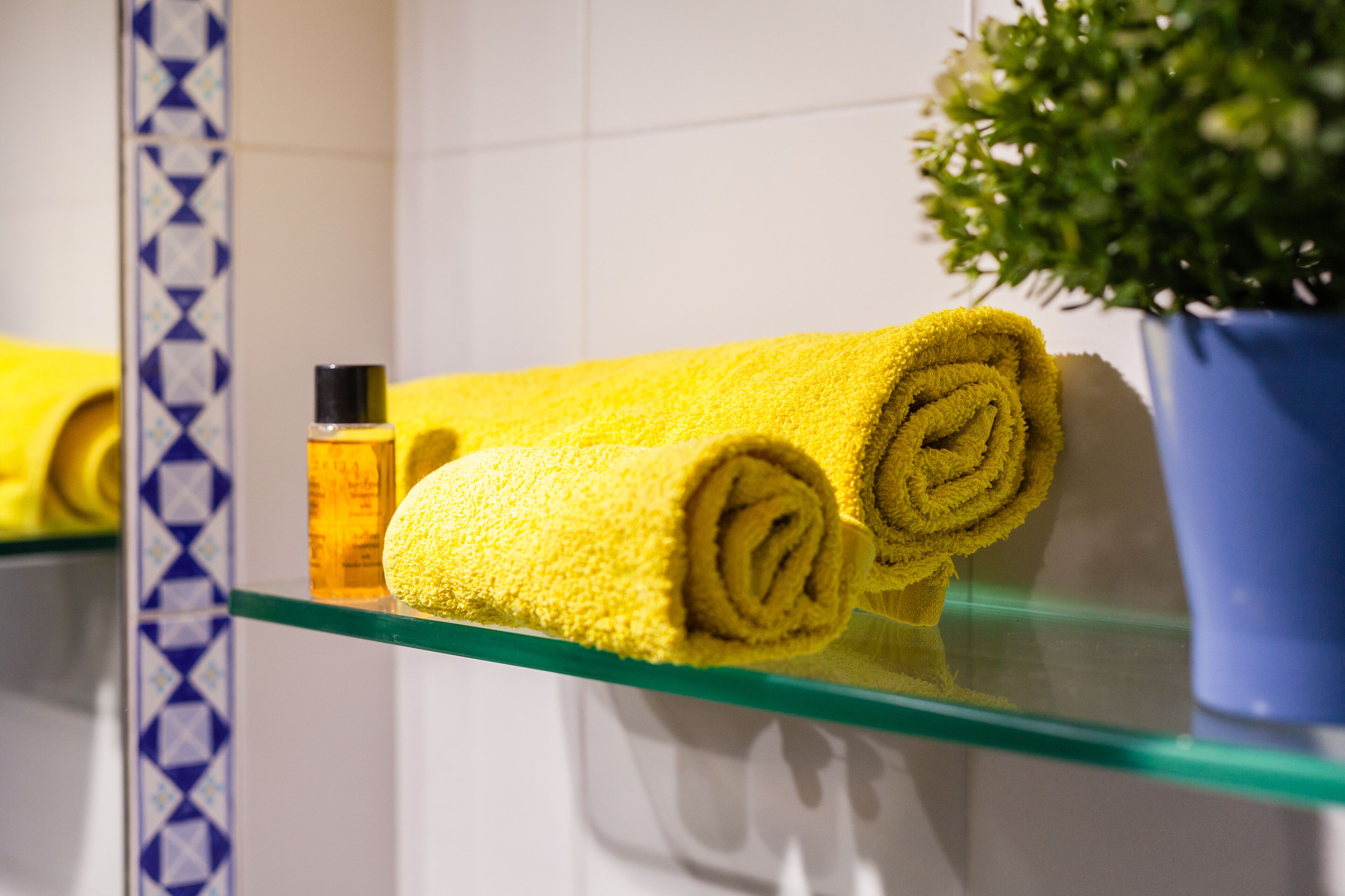 Un bagno con ripiano in vetro che contiene asciugamani gialli arrotolati, una lozione e una pianta in vaso.