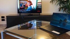 A television displaying a movie with Italian subtitles, a laptop on a glass table, and a potted plant beside the TV. - Sea View 2bed Apartment overlooking the Gulf with Private Terrace (Napoli)