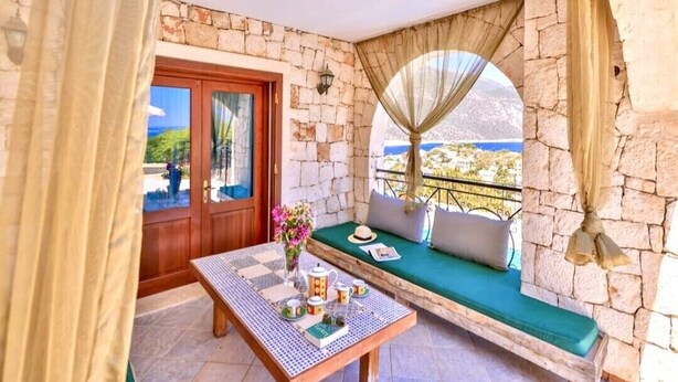 Un balcón acogedor con un muro de piedra, una puerta de madera, un banco acolchado y una mesa con un jarrón de flores.