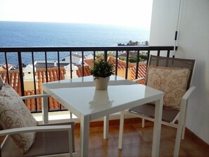 A balcony with a white table and two chairs, a potted plant, and a view of the sea and buildings.