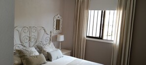 A bedroom with a bed, pillows, a nightstand, a lamp, a mirror, and a window with curtains.