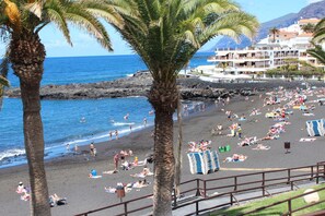 A beach with palm trees, a rocky shoreline, and a building with balconies overlooking the sea.
