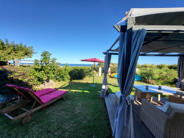 A covered outdoor seating area with a view of the sea, a wooden bench, and a table with cushions.