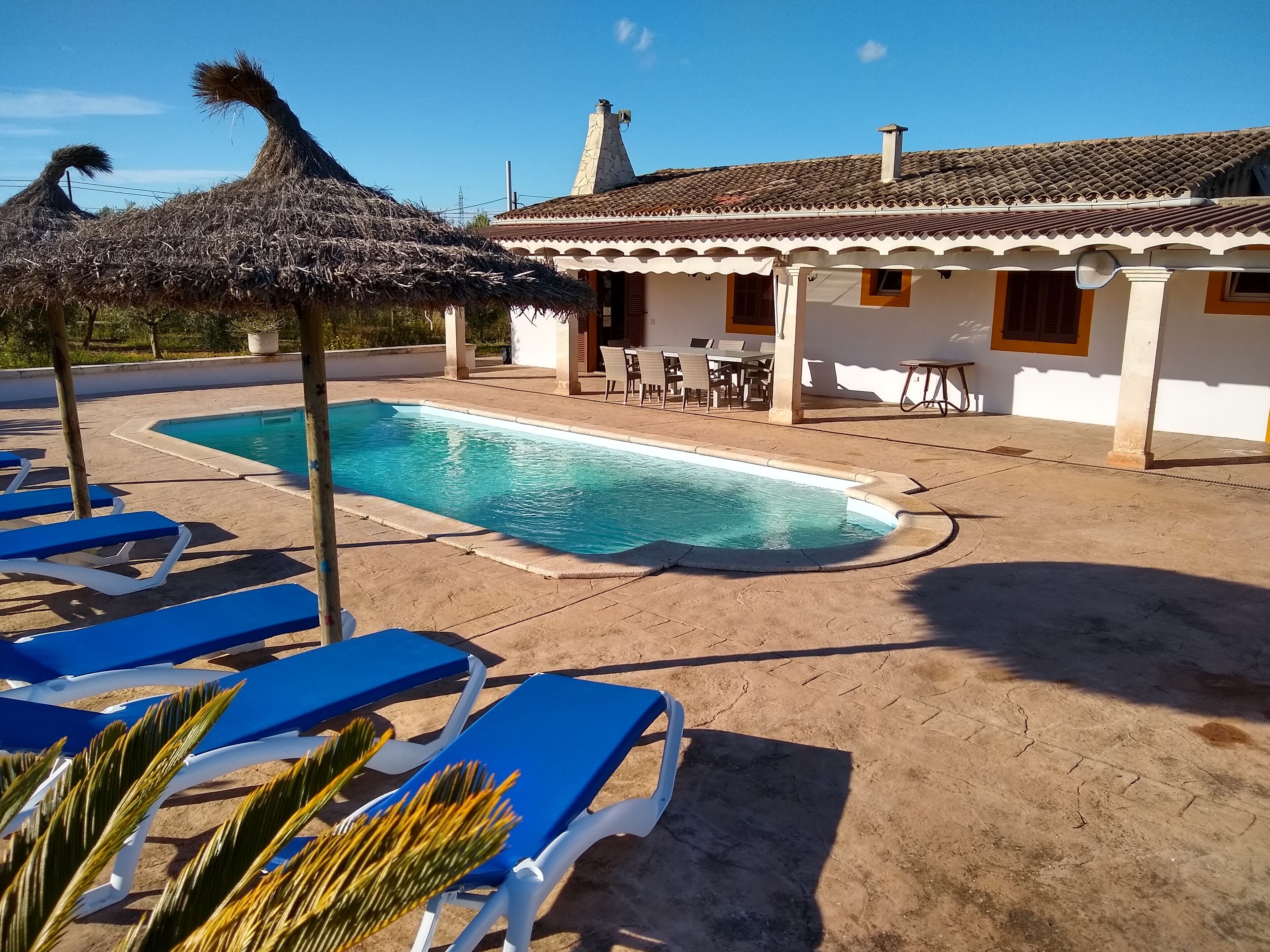 A swimming pool with blue lounge chairs, a thatched umbrella, and a building with a tiled roof and white walls.