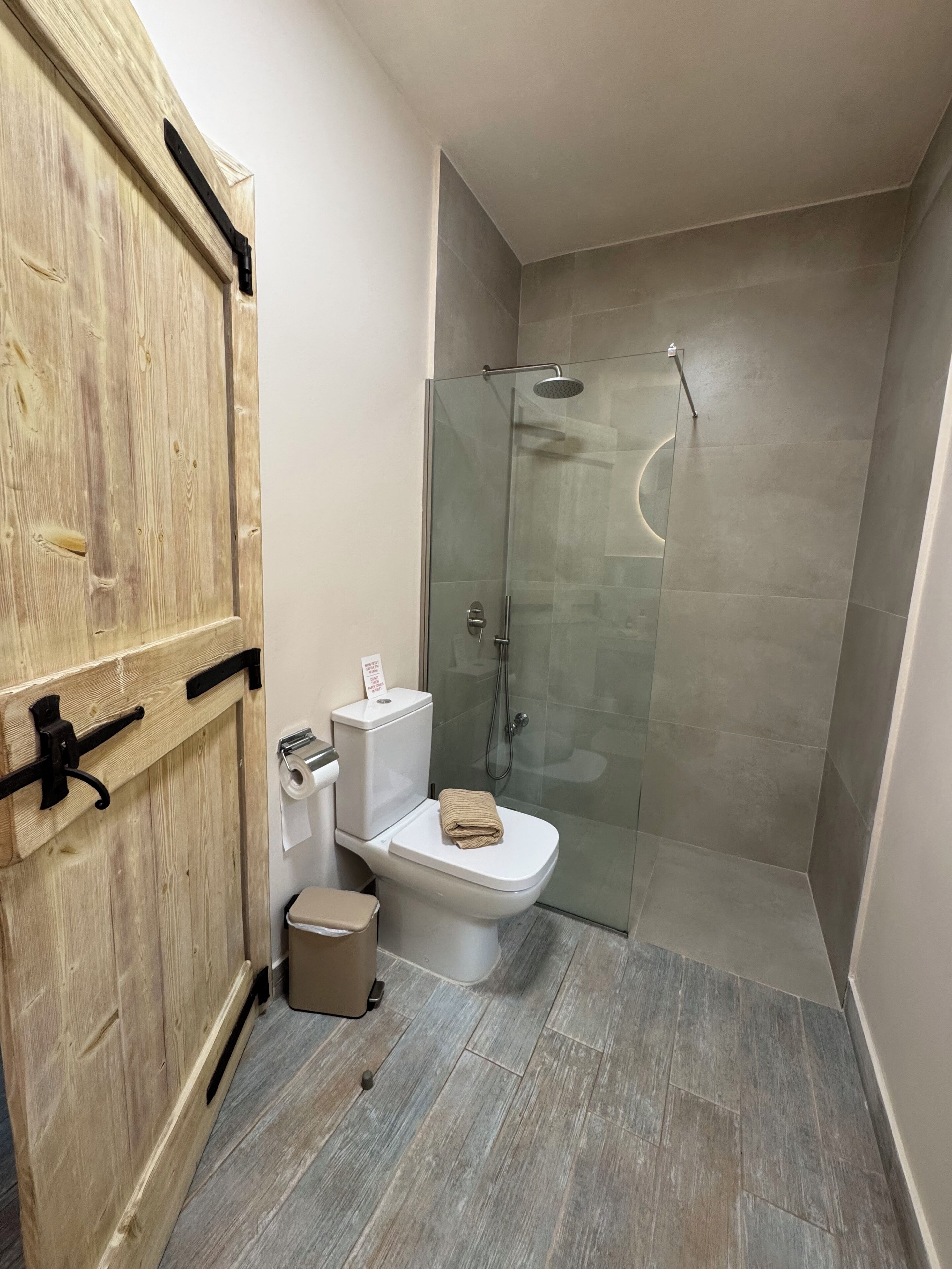 A bathroom with a wooden door, a toilet, a glass shower enclosure, and a small trash bin.