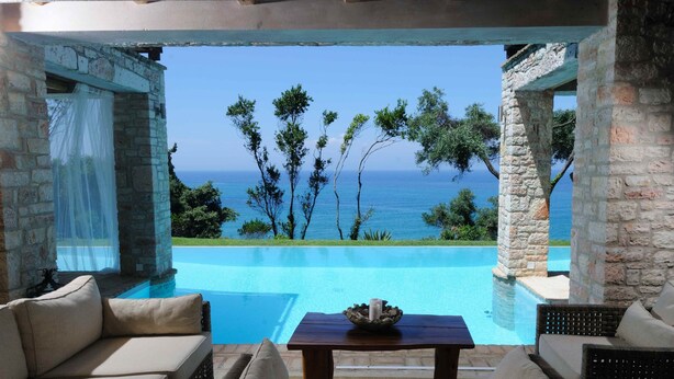 Zona junto a la piscina con vista al mar, pilares de piedra y mobiliario de mimbre.