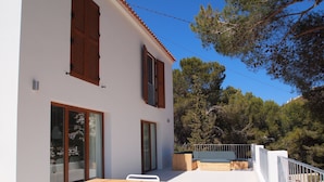 Une maison moderne à deux étages, dotée d’une terrasse en bois, de murs blancs et offrant une vue sur une colline boisée.