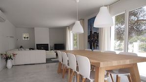 Un salon moderne avec un coin repas, comprenant une table en bois et des chaises blanches, un canapé, un téléviseur à écran plat et de grandes fenêtres offrant une vue sur le paysage urbain.