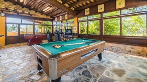 A pool table with a wooden frame and a stone floor, surrounded by windows with a view of greenery.