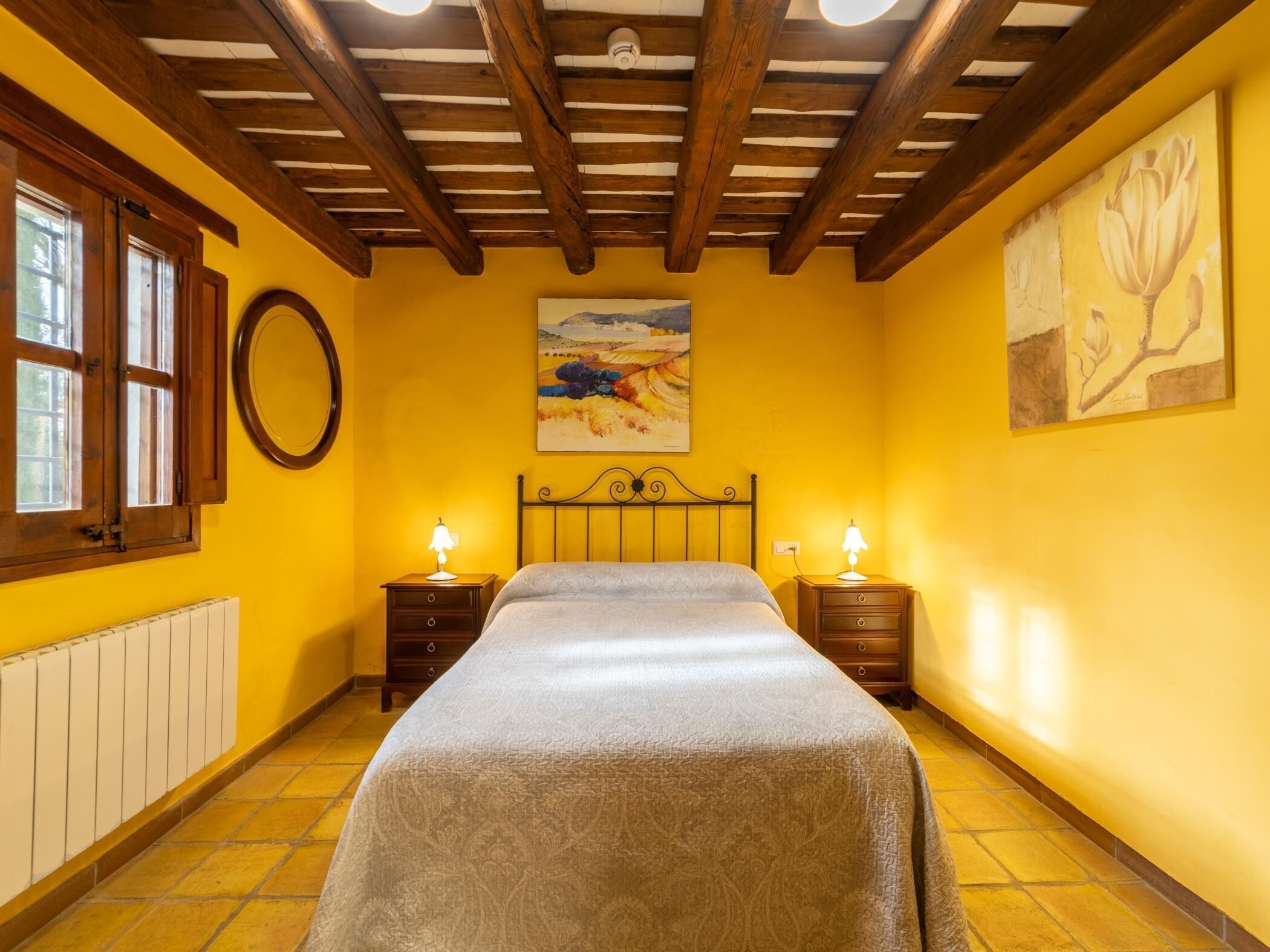 A bedroom with a bed, two bedside tables with lamps, a radiator, a window with wooden shutters, and a painting on the wall.