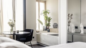 Een moderne hotelkamer met een bed, een ronde tafel met stoelen, een potplant en een zithoekje bij het raam.