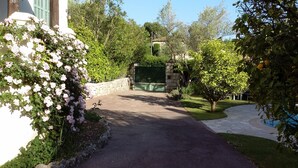 A paved driveway leading to a gated entrance, surrounded by lush greenery and flowering bushes. - Air-Conditioned Cottage With Pool in Vence. (Vence)