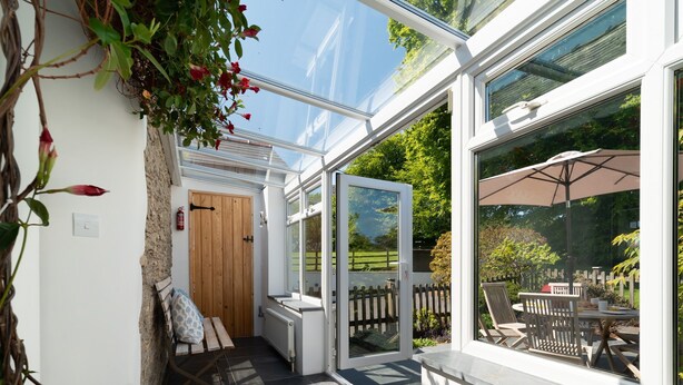 Een zonnig terras met een houten bank, een glazen deur naar een tuin, en uitzicht op een houten hek en tuinmeubilair.