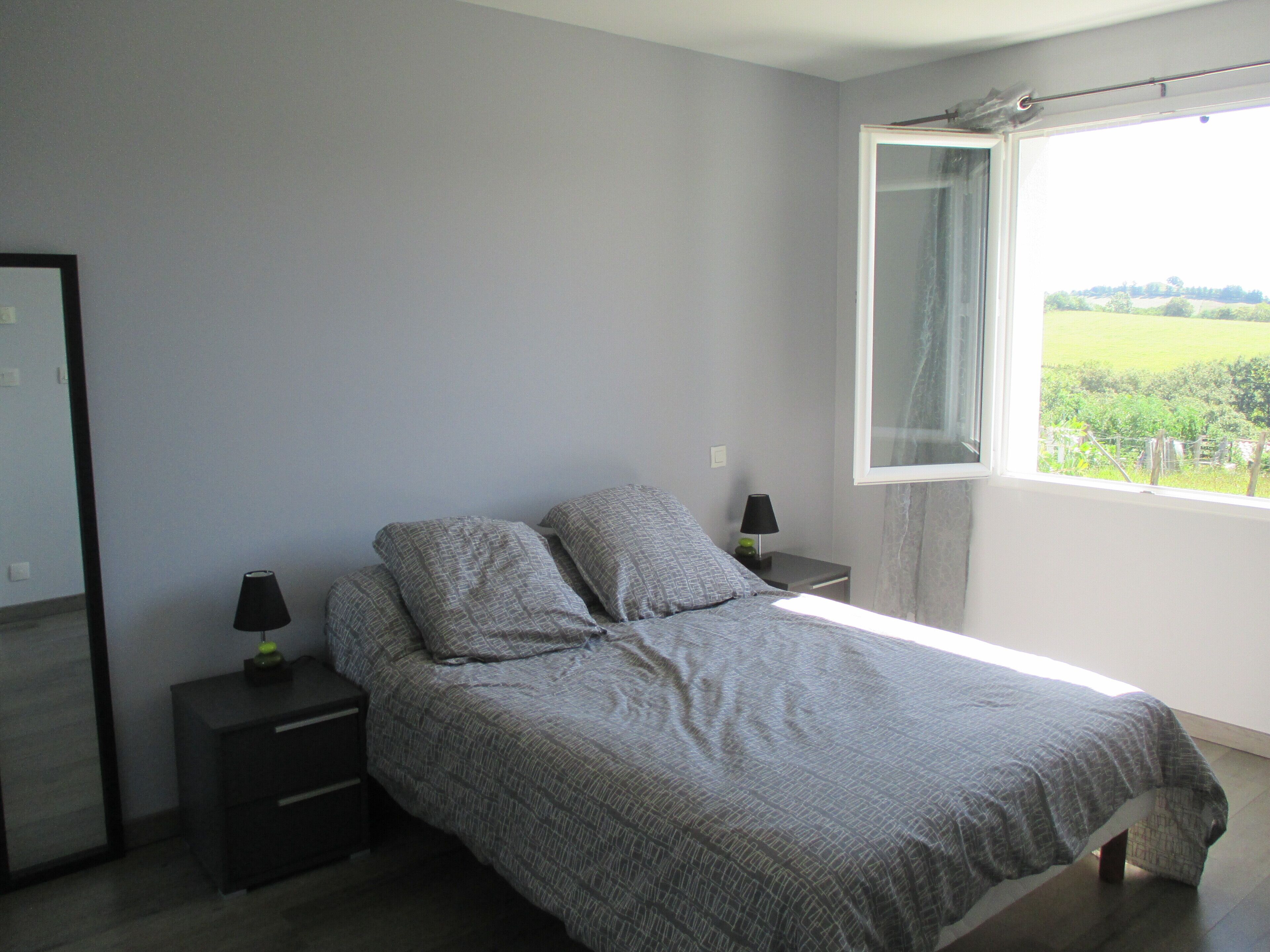 A bedroom with a bed, two bedside tables, a mirror, and a window with a view of greenery.