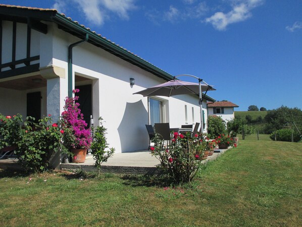 Un edifici blanc amb teulada verda, un pati amb mobles d'exterior i un jardí ple de flors roses.