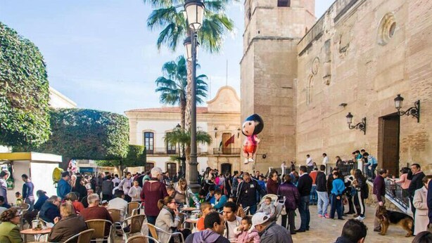 Un espacio al aire libre concurrido, personas sentadas en mesas, un edificio de arquitectura de piedra y palmeras.