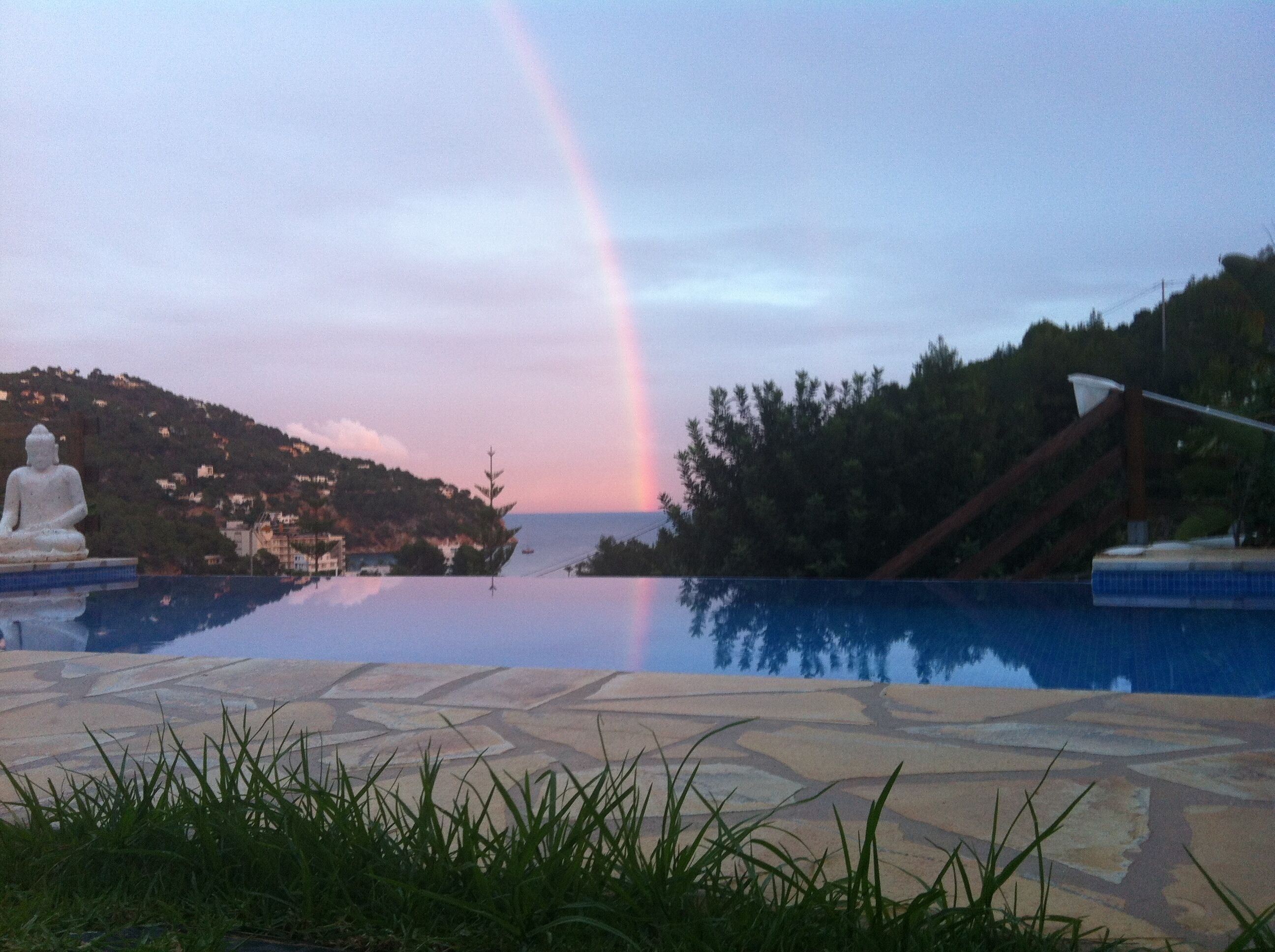 A serene pool area with a Buddha statue, a rainbow, and a view of a hillside with buildings.