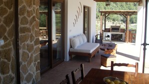 A cozy outdoor seating area with a stone wall, a cushioned sofa, and a dining table with a basket of fruit.
