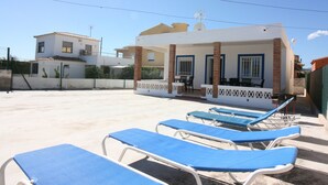 Zona de piscina con sillones reclinables azules y un área de estar cubierta con mesa y sillas.
