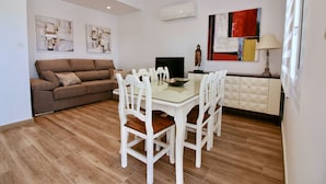 Una sala de estar moderna con un sofá marrón, una mesa de comedor de vidrio con sillas blancas, un televisor de pantalla plana y arte abstracto en la pared.