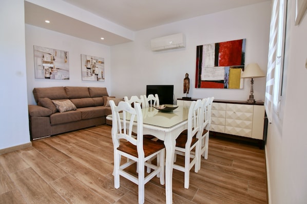 A modern living room with a brown sofa, a glass-top dining table with white chairs, a flat-screen TV, and abstract wall art.