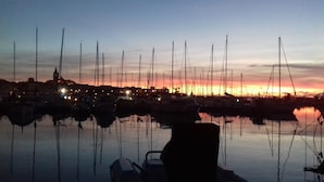 Un port de plaisance au crépuscule, avec de nombreux voiliers amarrés, des eaux calmes reflétant les bateaux et, au loin, la silhouette d’une ville.