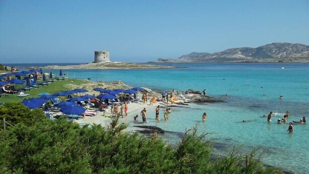 Une plage à l’eau turquoise et limpide, une structure en arrière-plan qui ressemble à un château, et des gens qui nagent et prennent des bains de soleil.