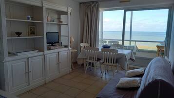 A room with a TV on a cabinet, a dining table with chairs, a sofa, and a view of the beach.