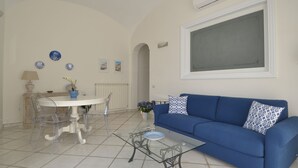 A living room with a blue sofa, a glass coffee table, a white dining table with chairs, and a potted plant. - L'Ancora - Bright Sea front, Self Catering Apartment (Minori)