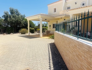 A covered outdoor seating area with hexagonal paving, a wooden fence, and a building with a balcony.