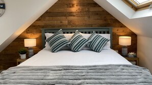 A bedroom with a sloped ceiling, a wooden headboard, a large bed with striped pillows, two bedside lamps, and a skylight. - Beautiful Seaside Cottage 1 minute from the beach - The Tides (Camber)