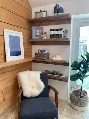 A cozy room with wooden walls, a framed picture, a chair with a fluffy pillow, and shelves with books, decorative items, and a potted plant.