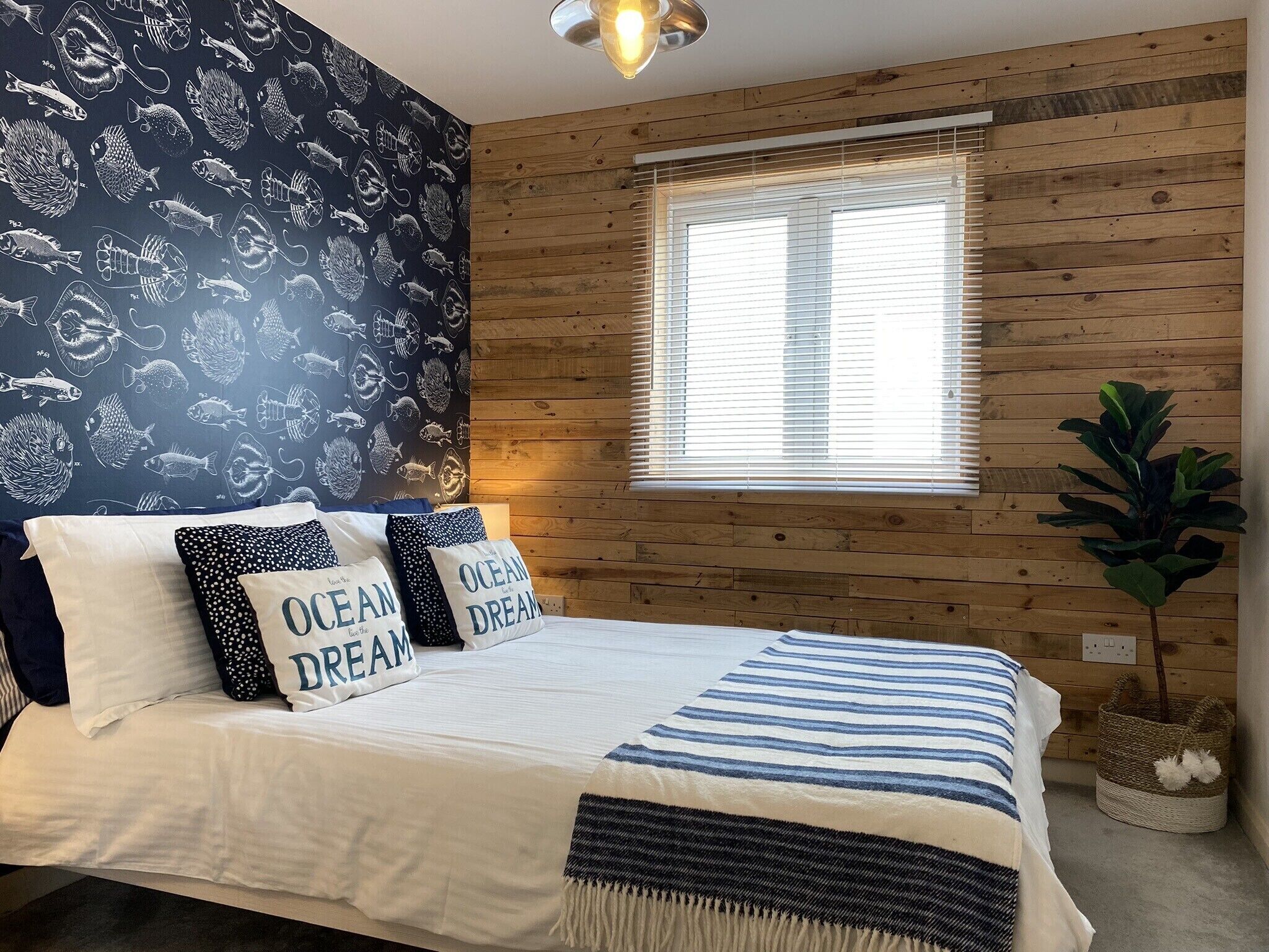 Une chambre avec un mur en bois, une fenêtre avec des stores, un lit avec une literie à rayures et deux oreillers ornés de la mention « OCEAN DREAM ».