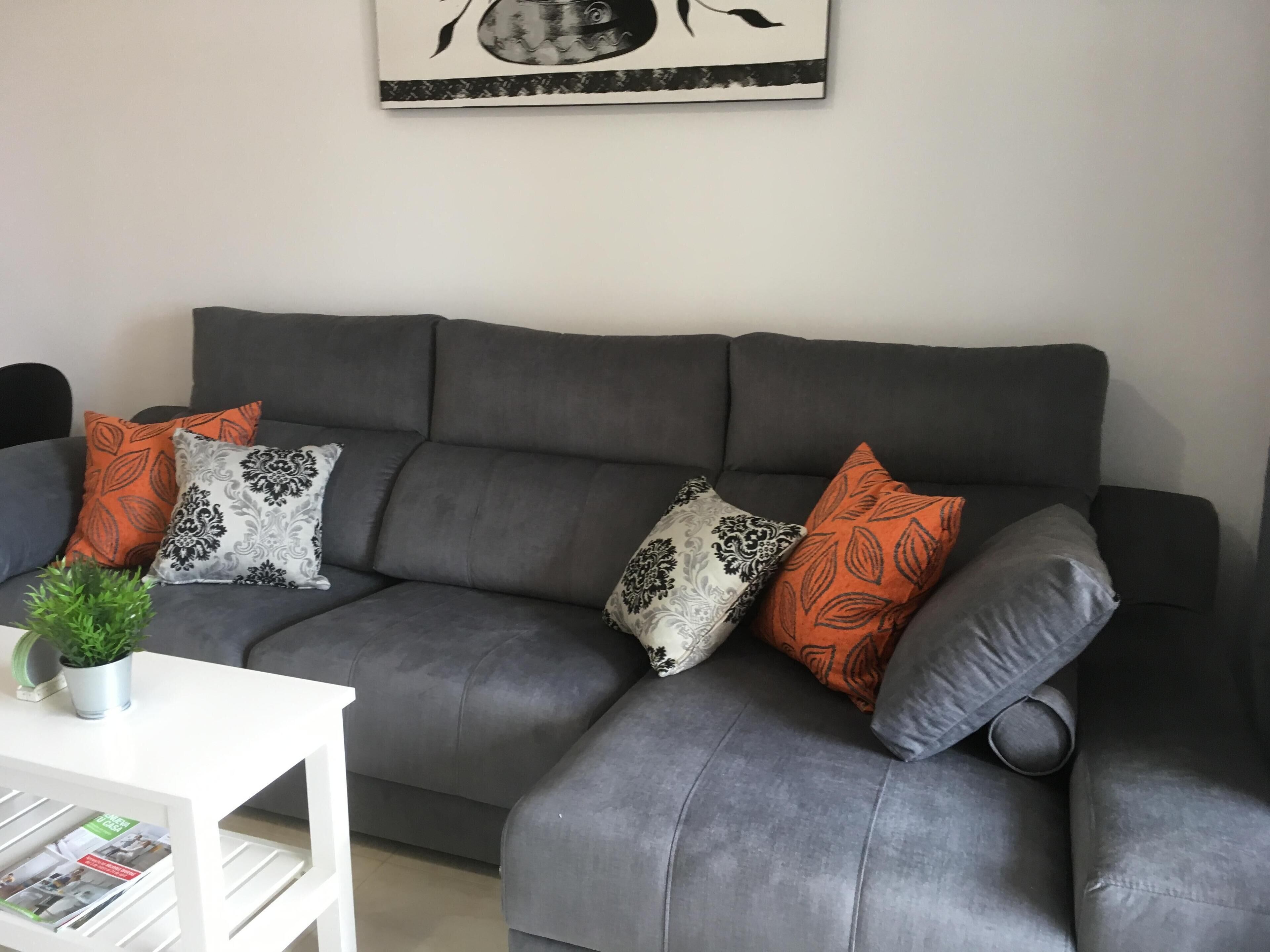 A grey sofa with patterned cushions, a small potted plant, and a white side table with magazines.