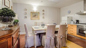 Un comedor con una mesa dispuesta para cuatro personas, rodeada de sillas. Se observa una zona de cocina con electrodomésticos y armarios.
