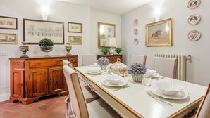 Un comedor con una larga mesa dispuesta para cuatro, rodeada de sillas. A la izquierda, un aparador de madera y, a la derecha, un radiador. Las paredes están decoradas con cuadros enmarcados y espejos.