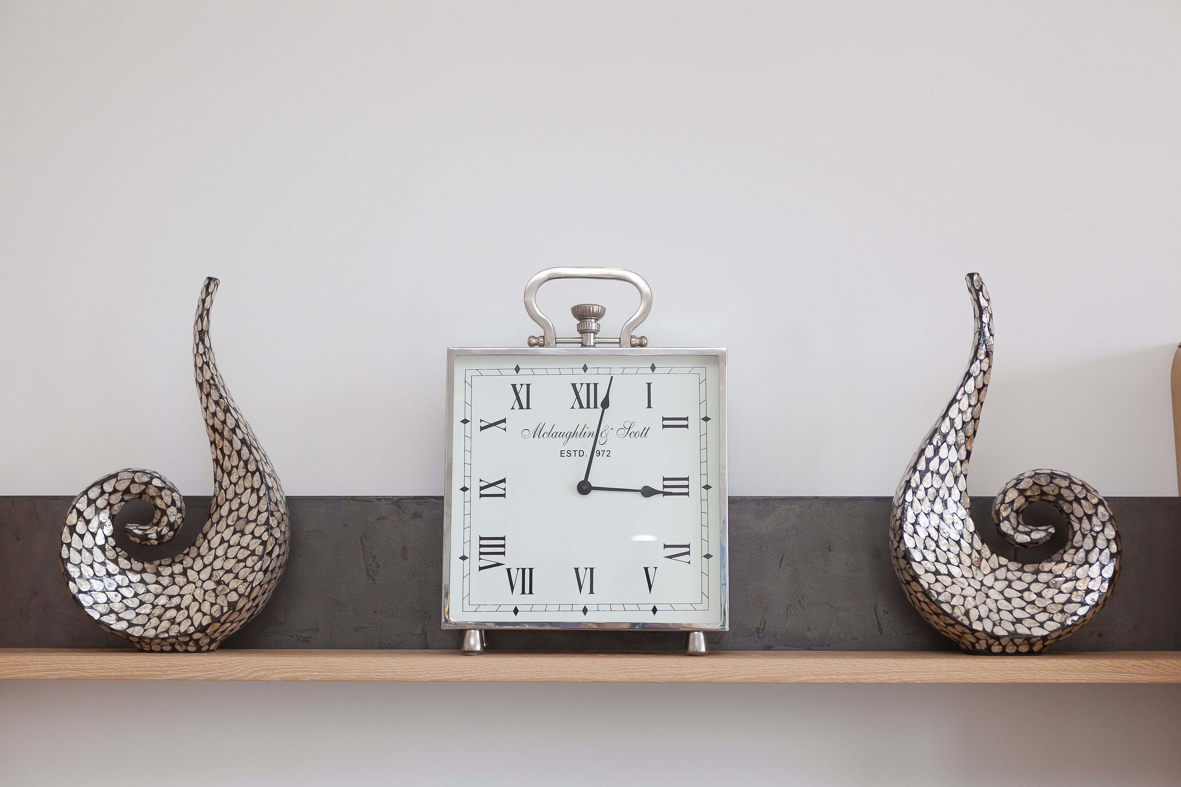 Reloj de pared con acabado metálico y esfera blanca, flanqueado por dos objetos decorativos con diseño de hojas a cada lado.