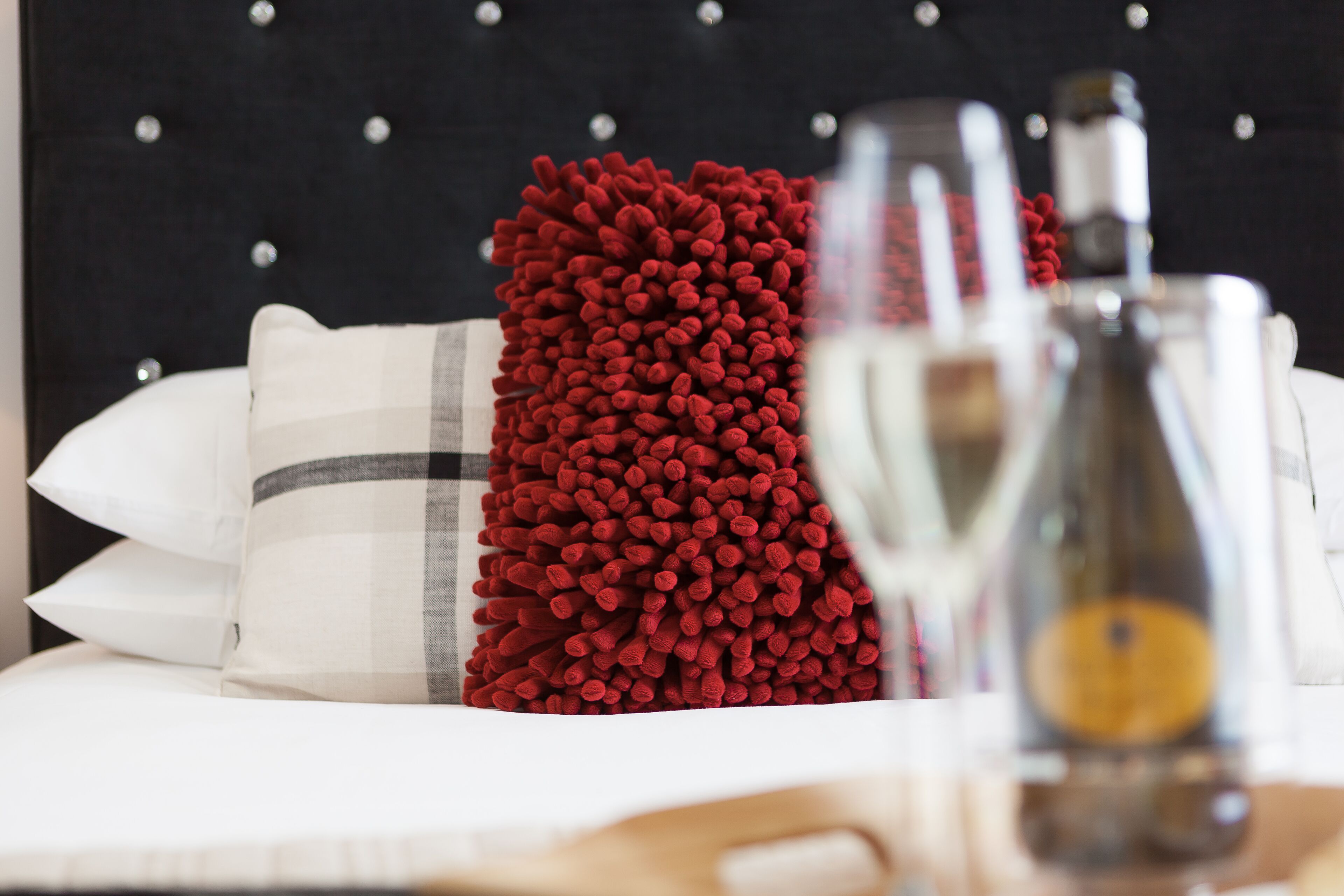 Un lit avec des oreillers blancs et gris, un oreiller rouge à texture, et une bouteille en verre transparent contenant un objet jaune.