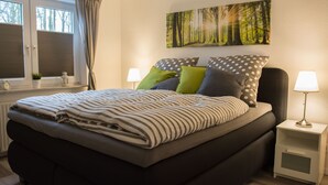 Une chambre moderne avec un grand lit, deux tables de chevet, une fenêtre avec des rideaux et une œuvre d’art murale sur le thème de la forêt.