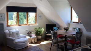 Et hyggeligt loftsværelse med en hvid sofa, et spisebord med stole, et fjernsyn og et stort vindue med udsigt over grønne områder.