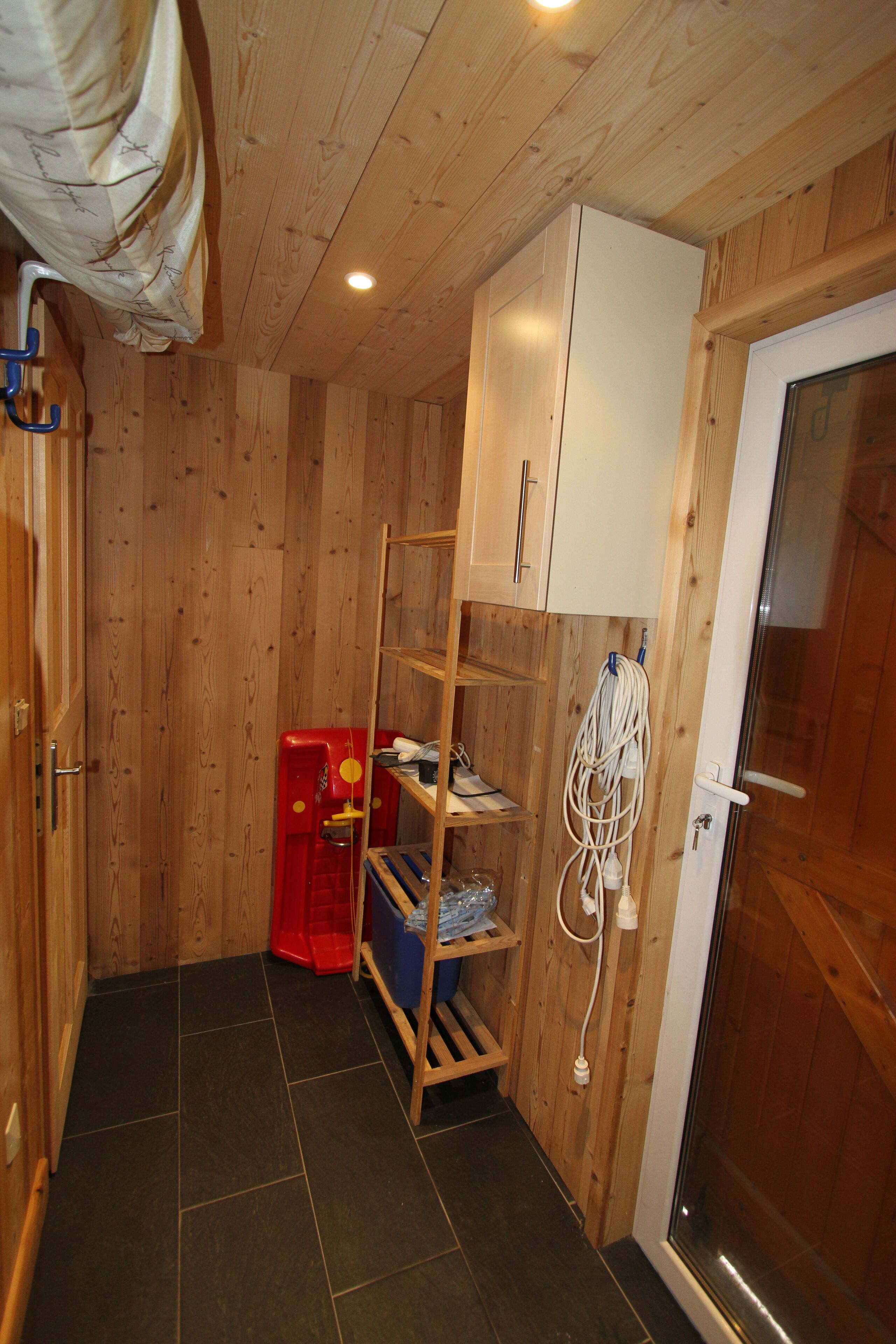 A compact room with wooden walls, a tiled floor, a white door, and a small shelving unit with various items.