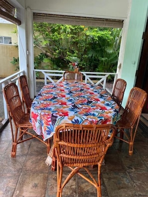 Un espace extérieur abrité, aménagé pour les repas, avec une table ronde et six chaises en osier, entouré d’une balustrade blanche et d’une végétation luxuriante en arrière-plan.