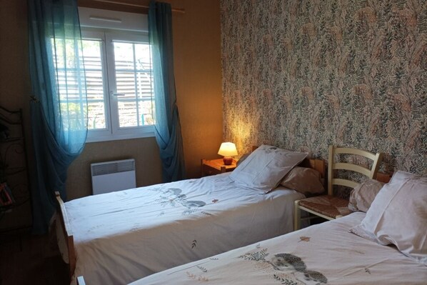 A bedroom with two beds, a window with curtains, a bedside lamp, and a radiator.