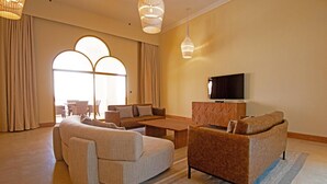 A spacious living room with a large sofa set, a coffee table, and a flat-screen TV on a wooden cabinet. The room has large windows with curtains and a chandelier. - HOTEL (Abu Dhabi)