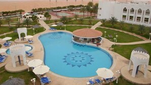 A swimming pool with a geometric design, surrounded by lounge chairs and umbrellas, with a view of the sea and a building in the background. - HOTEL (Abu Dhabi)