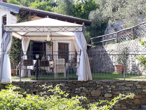 Uma área de estar coberta ao ar livre, com parede de pedra, guarda-corpo de metal e pérgola.
