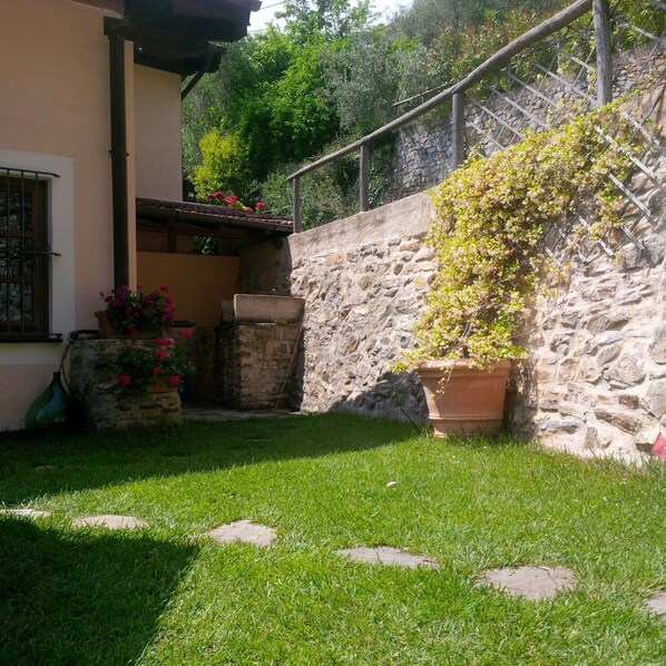 Un muro de piedra con una planta en maceta, un área con césped y piedras para pisar, y un edificio con ventana y macetas con flores.