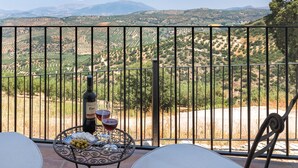 A balcony with a table set for two, featuring a bottle of wine, two glasses, and a plate of olives. The balcony overlooks a vineyard and distant mountains.
