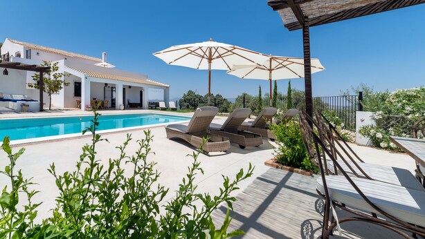 A poolside area with lounge chairs and umbrellas, a clear blue sky, and a white building with a tiled roof.