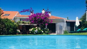 En swimmingpool med en klar blå vandoverflade, omgivet af et hus med et tagtækket tag, en have med blomstrende planter og et hvidt hegn.